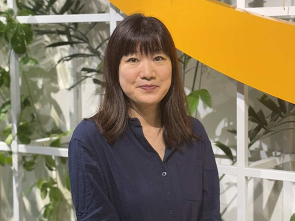 A woman wearing a navy shirt stands in front of a background with indoor plants and a white trellis, with an orange structure overhead.