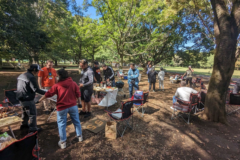 A group of people enjoying a barbecue gathering in a park, surrounded by trees and outdoor seating.