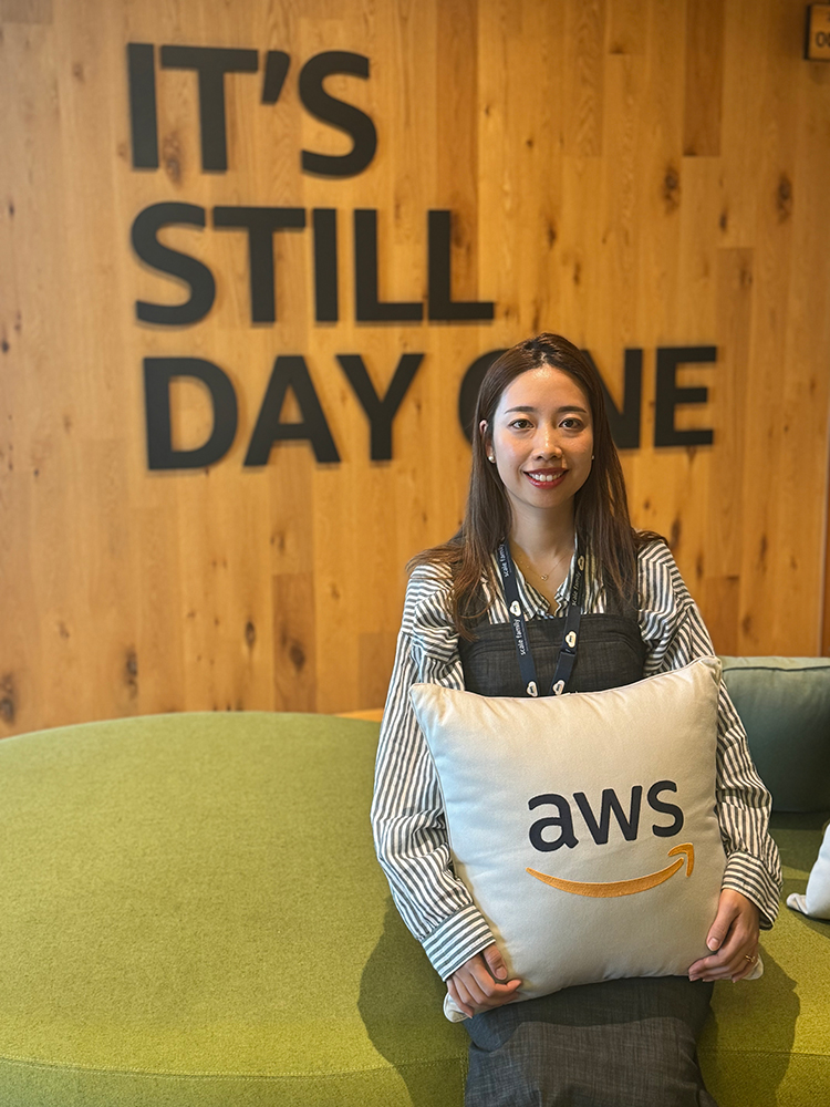 A person sits in an AWS office holding a pillow with the AWS logo. The wooden wall behind features the phrase 'IT'S STILL DAY ONE,' representing AWS corporate culture.