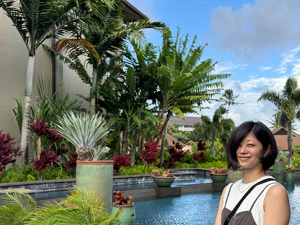 A woman smiling while standing poolside in a lush tropical garden, with palm trees and vibrant plants in the background under a blue sky.