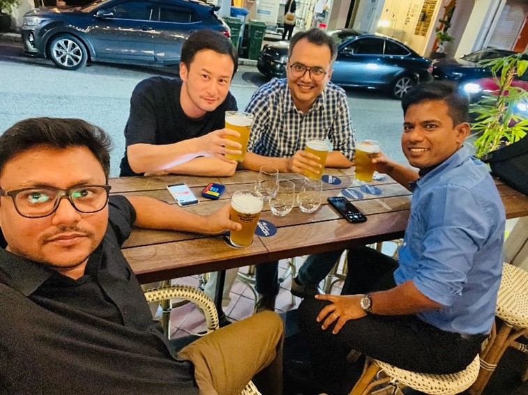 Four friends are sitting together at an outdoor bar, enjoying glasses of beer and smiling at the camera. Various items including mobile phones and empty glasses are on the wooden table. Cars and buildings are visible in the background, indicating a lively city street setting.
