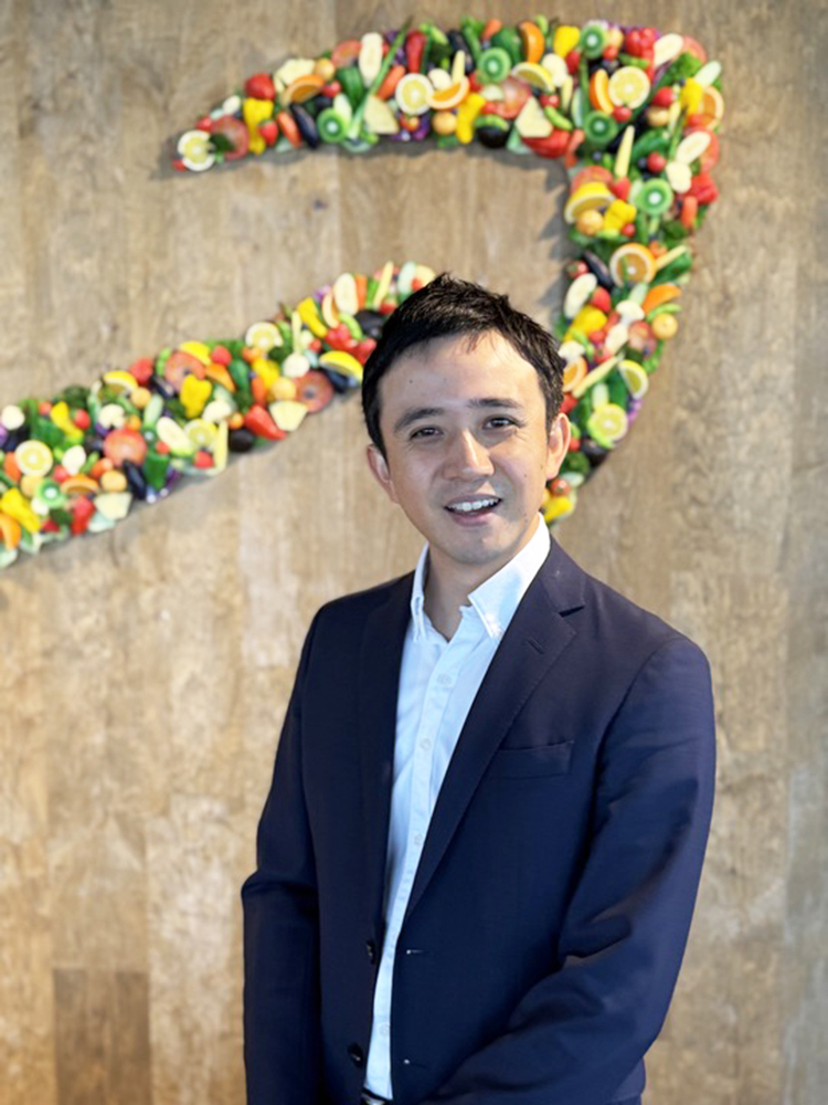 A man in a suit standing and smiling in front of a wall decorated with a colorful fruit logo.