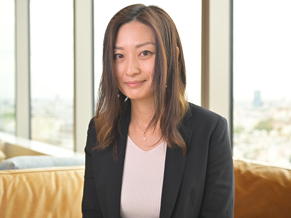 A businesswoman in casual office attire sits in front of large windows with an urban background, smiling slightly. She is wearing a black blazer and light top, with cityscape visible outside.