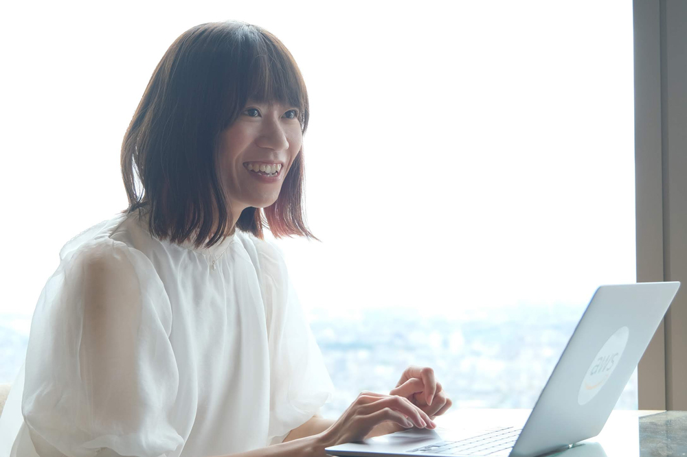 A woman in a white blouse is smiling while using a laptop that features the AWS logo, seated by a bright window.