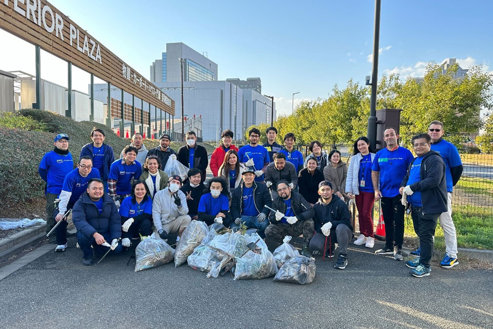 A group of volunteers pose together after a cleanup event at an exterior plaza, with several bags of collected trash in front of them. The event appears to be focused on community service and environmental responsibility.