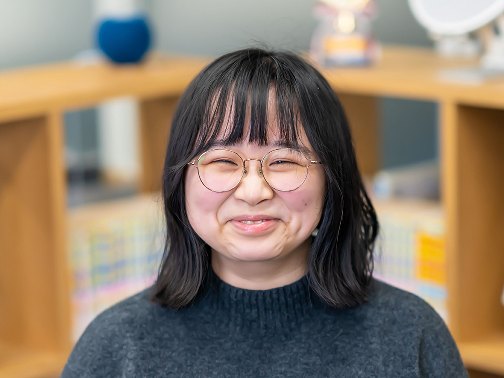 A smiling woman with glasses is standing indoors in front of a bookshelf background.