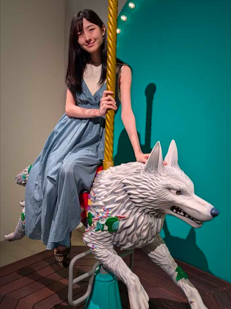 A woman in a blue dress is riding a carousel designed as a white wolf statue, indoors with colorful decor.
