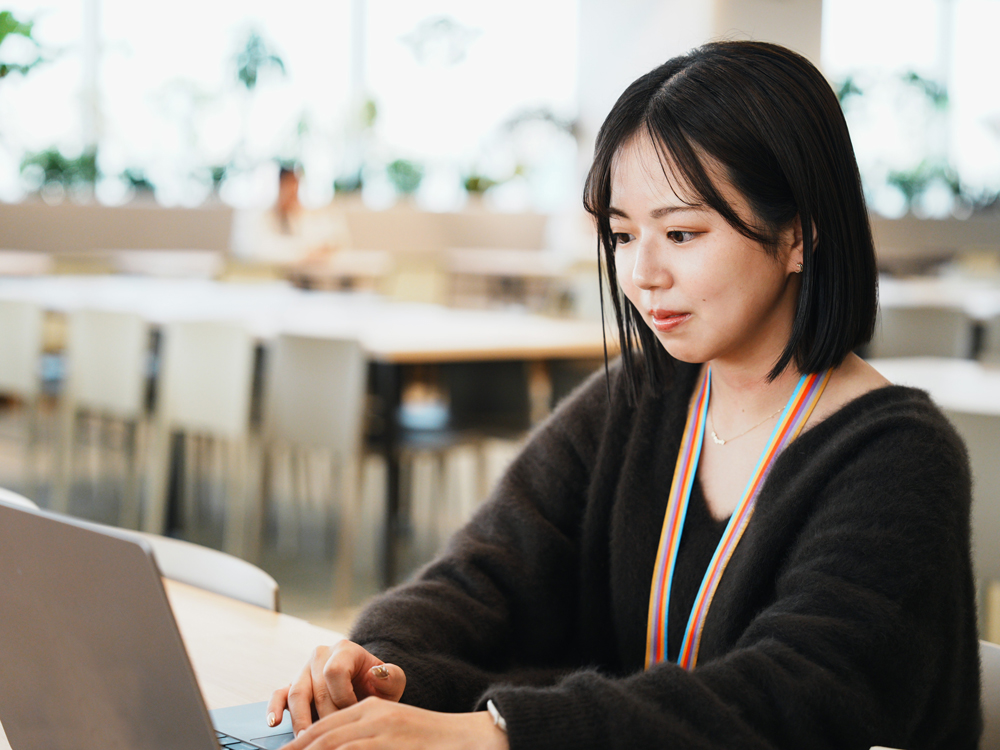 A woman working on a laptop in a modern, bright office environment with large windows and contemporary furniture.