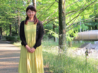 A woman in a yellow dress and black sweater standing in a sunny park with trees and greenery around her.