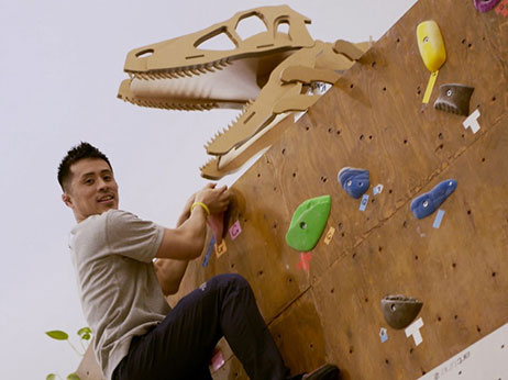 A man climbs an indoor climbing wall decorated with colorful holds and a large dinosaur head sculpture above the wall.