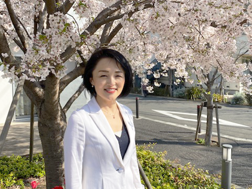 A woman wearing a white blazer stands under a cherry blossom tree in bloom on a sunny day.