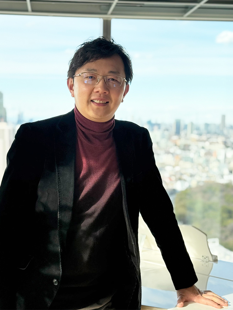 A man wearing glasses, a blazer, and a turtleneck stands by an office window with a city skyline view in the background.
