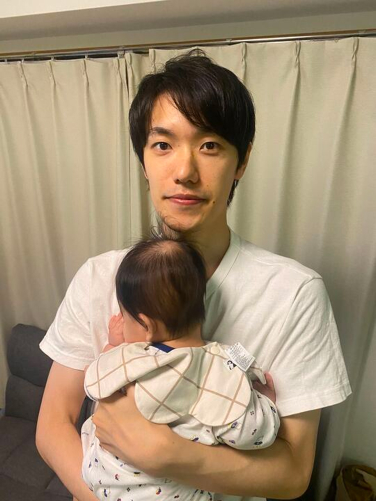 A man in a white t-shirt is gently holding a baby at home, standing in front of a curtain. The baby is resting its head on the man's chest, facing away from the camera.