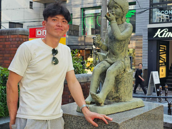 A man in a white t-shirt standing next to a child statue on a city street, with urban buildings and signs in the background.