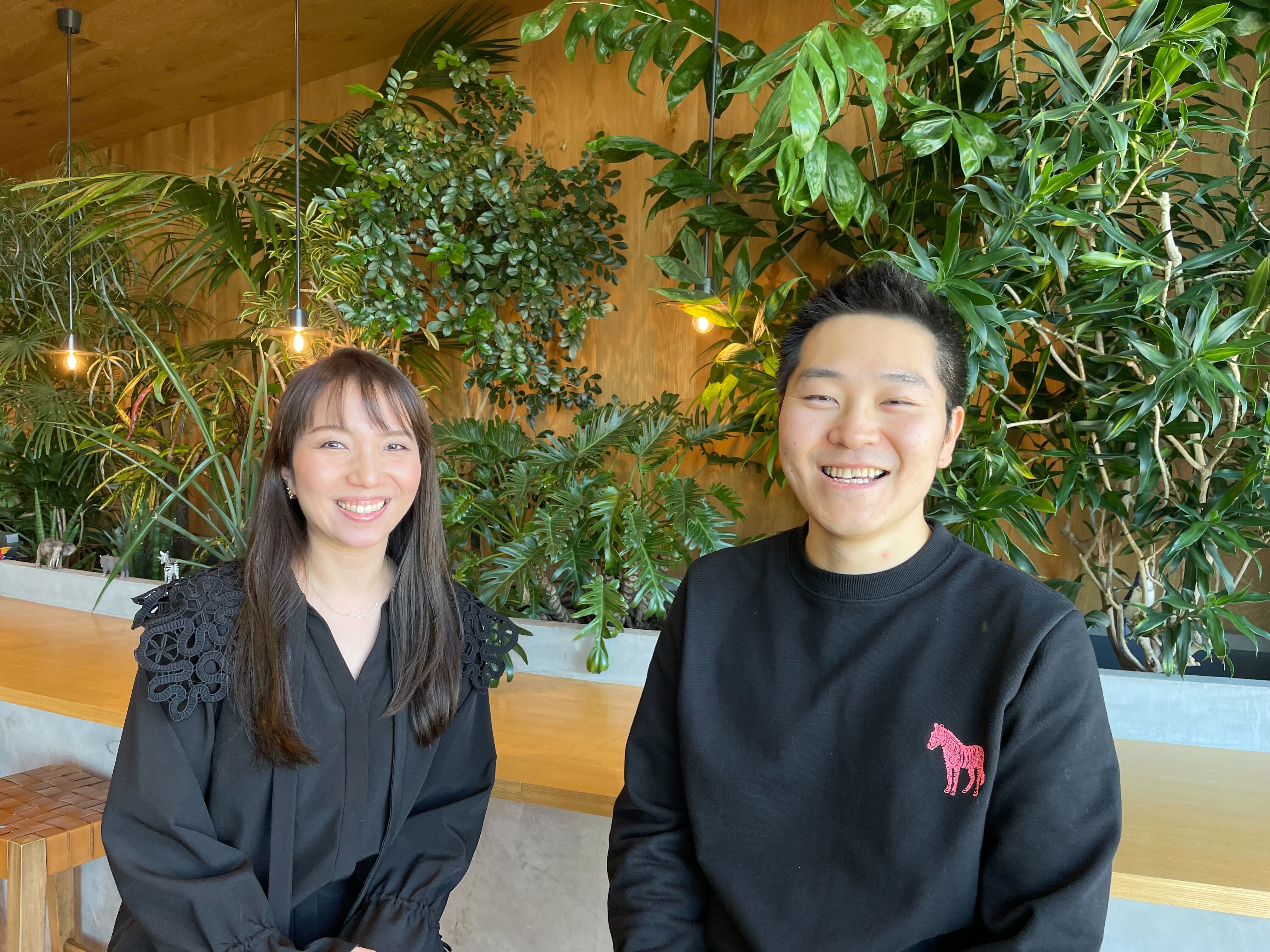 Two smiling coworkers sitting in a modern office space with lots of green plants and wooden decor in the background.
