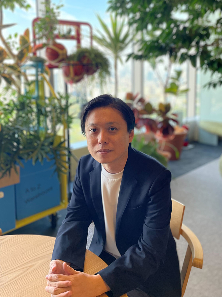A man wearing a black blazer and white shirt is sitting at a wooden table, with a variety of lush indoor plants in the background.