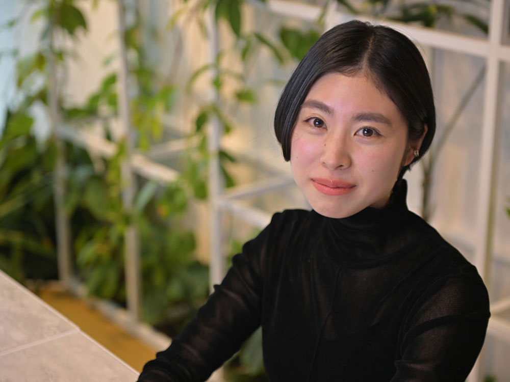 A woman in a black turtleneck sits at a table with a background of green indoor plants.