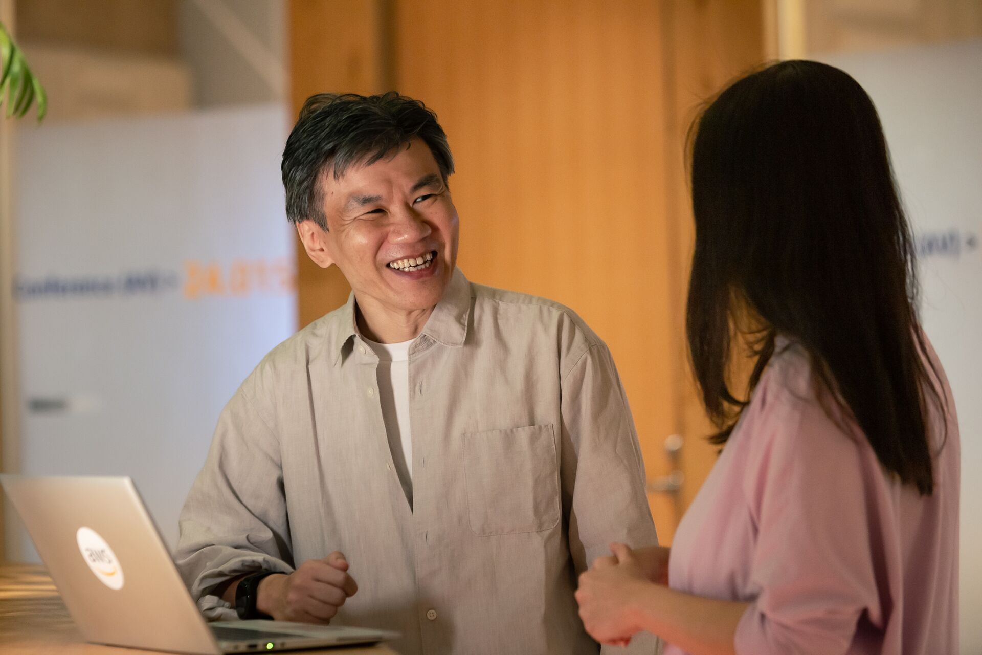 Two colleagues having a friendly conversation in an office environment, with an AWS-branded laptop on the desk.