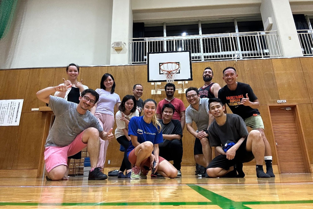 A group of people posing together on a gymnasium basketball court, smiling in front of a basketball hoop. The group appears to be a recreational or corporate basketball team enjoying their time indoors.