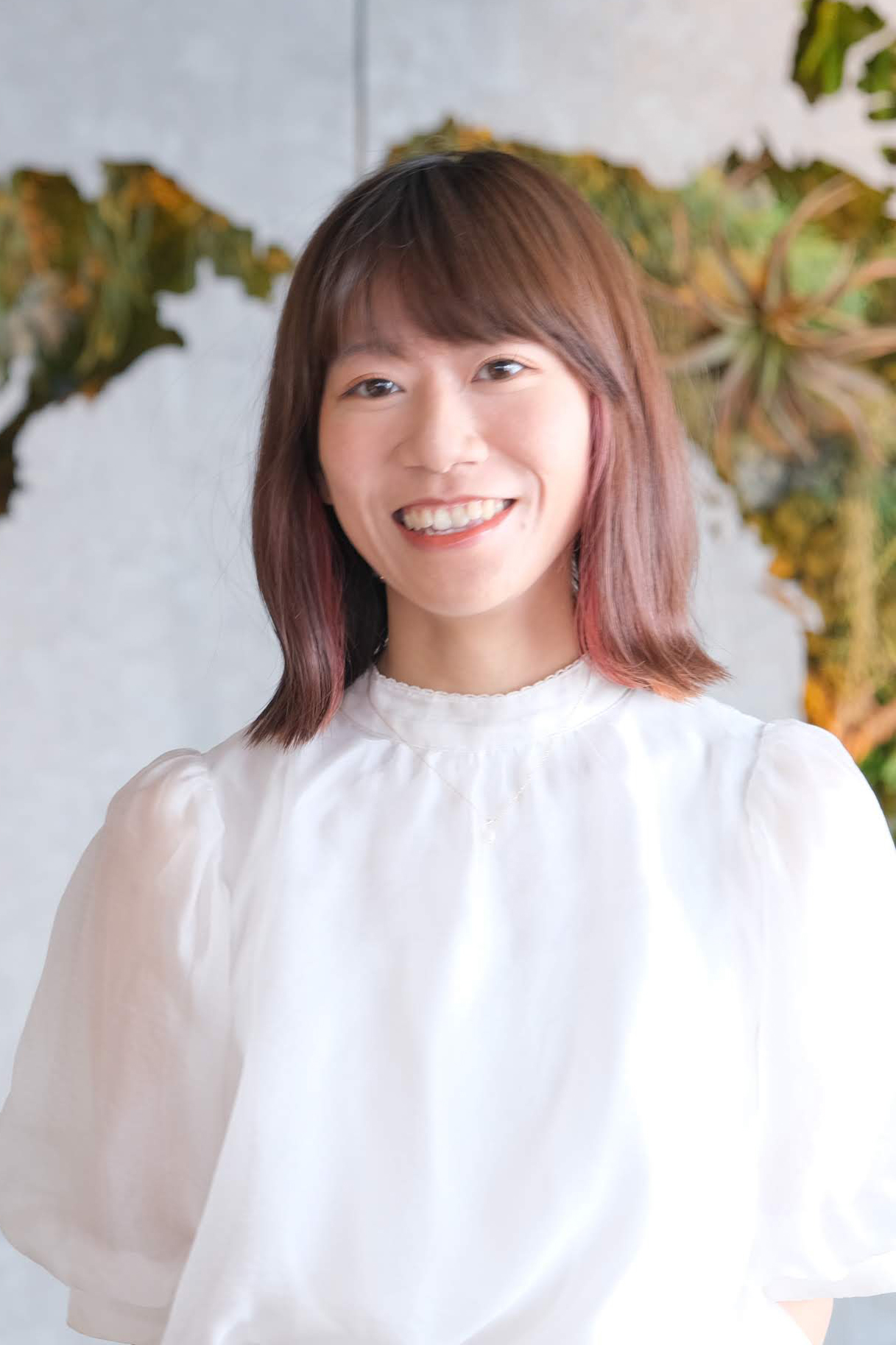 A portrait of a smiling woman wearing a white blouse, photographed indoors with a blurred background featuring green plants.