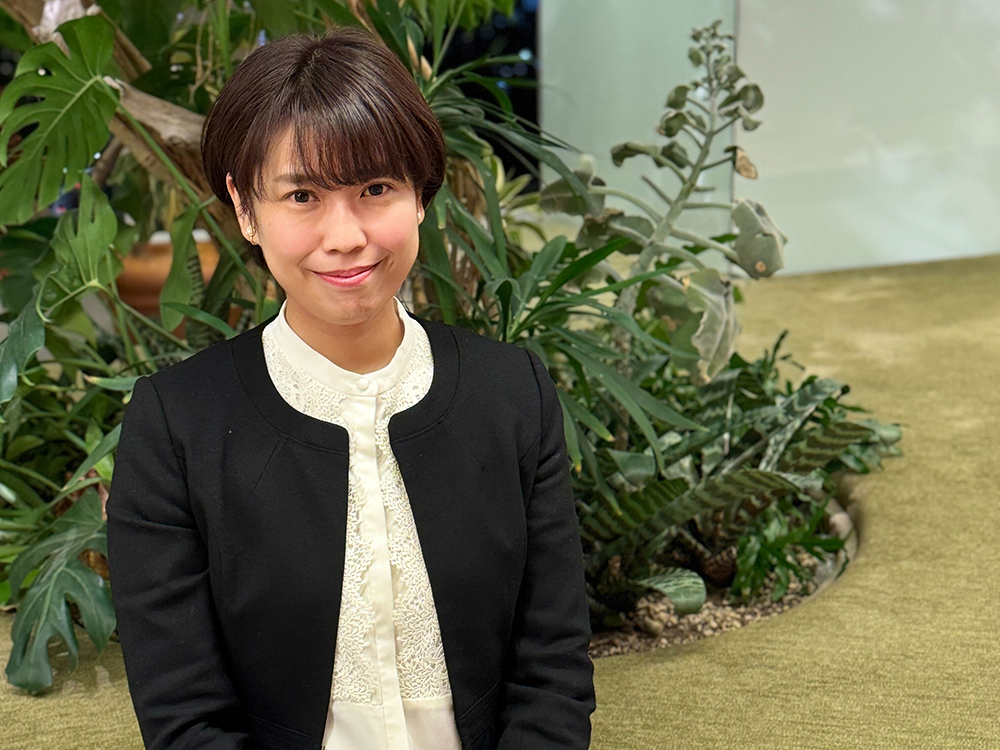 A woman wearing a black jacket and white blouse is sitting in front of an indoor garden area with various green plants. The setting suggests a modern, nature-inspired workspace or lounge.
