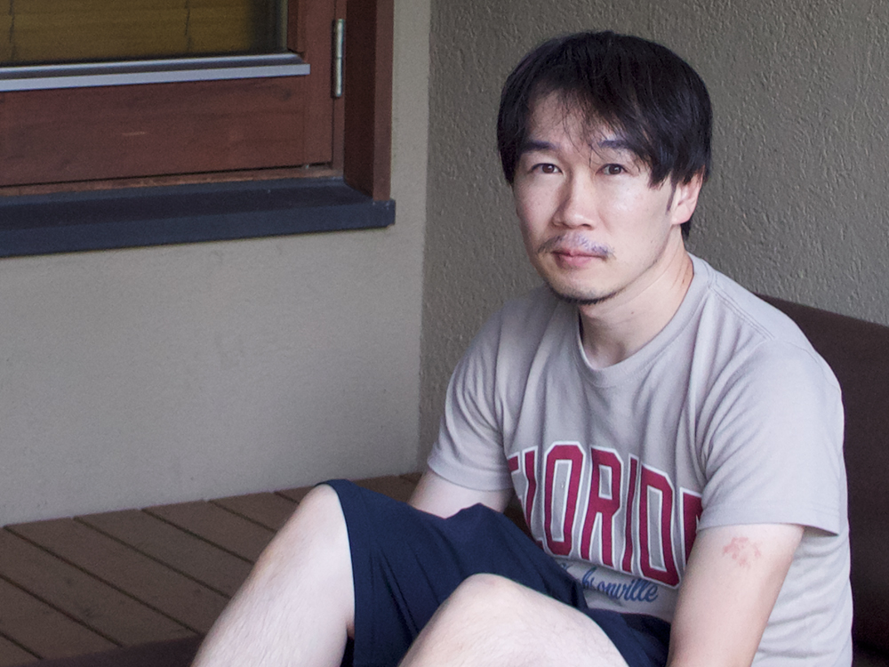 A man sitting casually indoors, wearing a Florida t-shirt and dark shorts, next to a window.