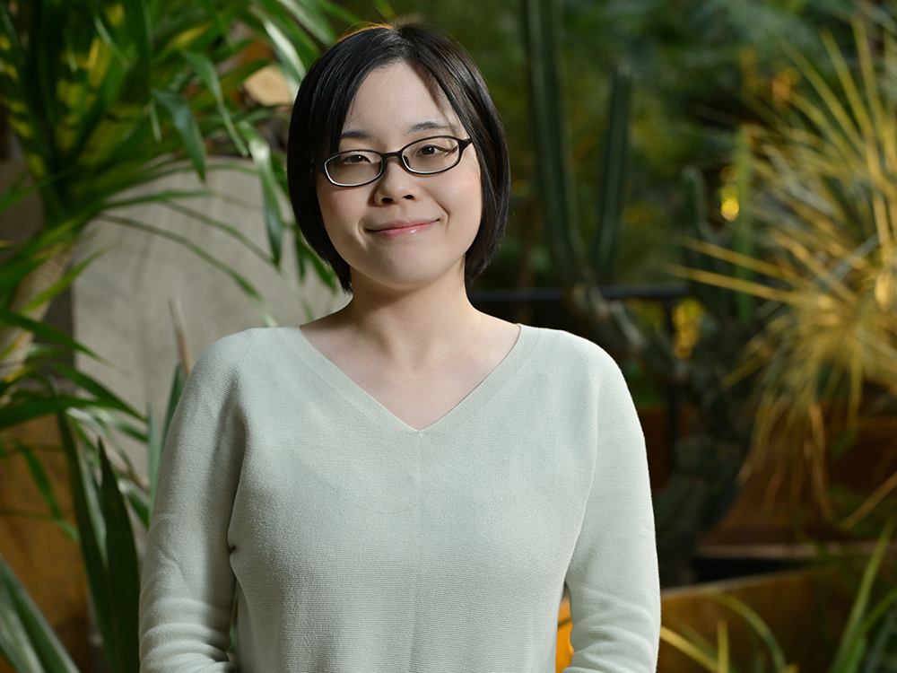 A woman wearing glasses stands in front of a background filled with lush indoor plants.