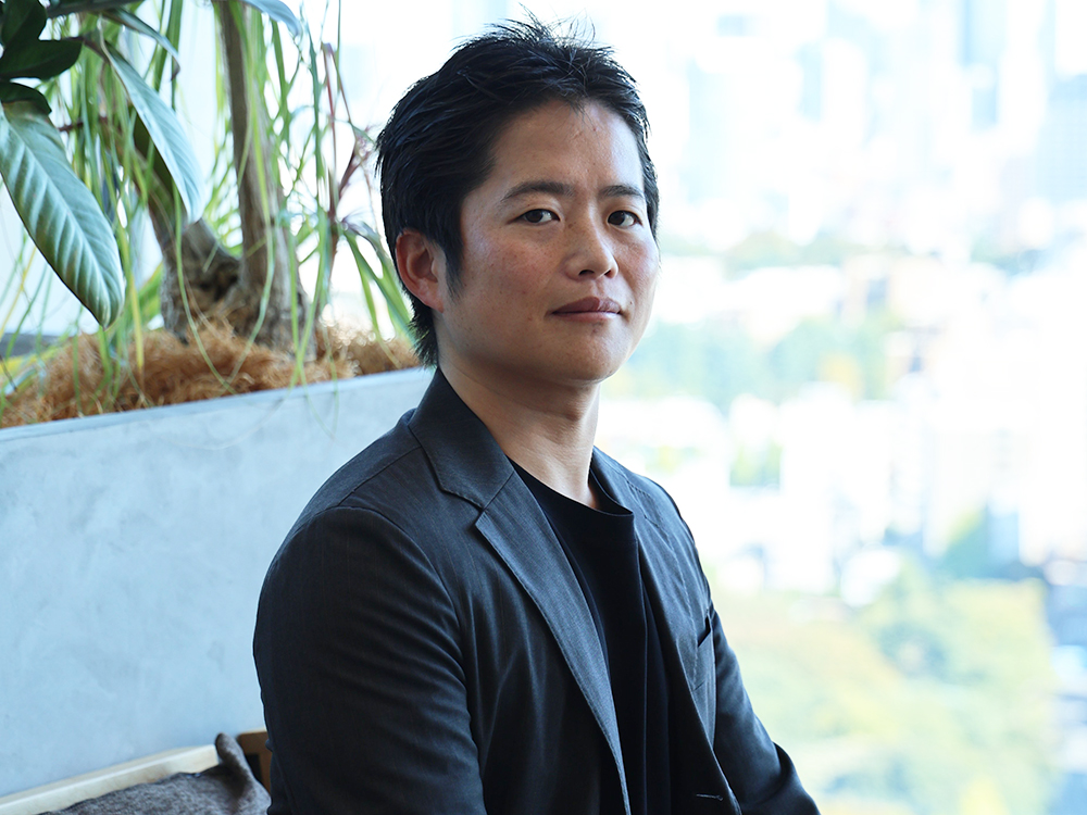 A businessman in a dark jacket sits indoors with green plants and a cityscape visible in the background.