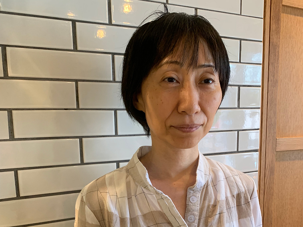 A woman standing in front of a white tiled wall, wearing a light-colored, patterned shirt.