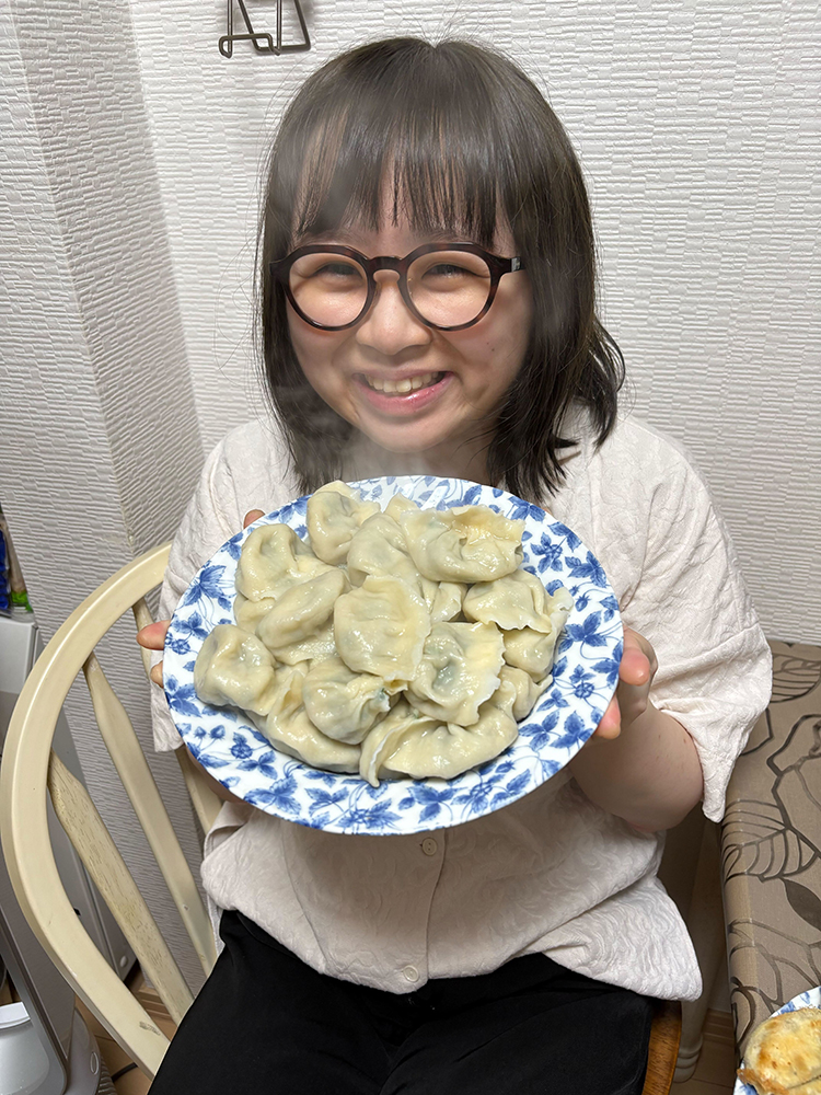 A smiling person with glasses holds a blue and white plate filled with steamed dumplings, sitting at a table indoors.
