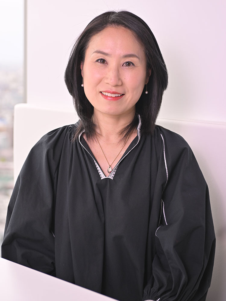 A woman with shoulder-length black hair is smiling while seated, wearing a black top. The setting is bright and modern, with a soft, neutral background.