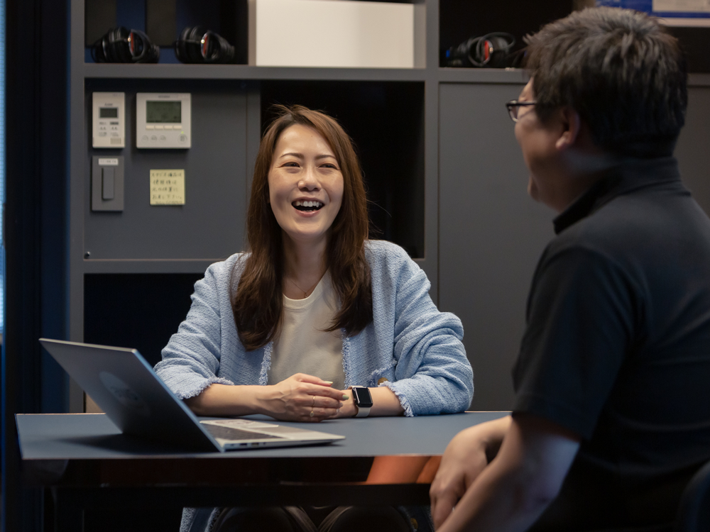 Two colleagues having a casual meeting in a modern office, sitting at a table with a laptop, smiling and conversing.