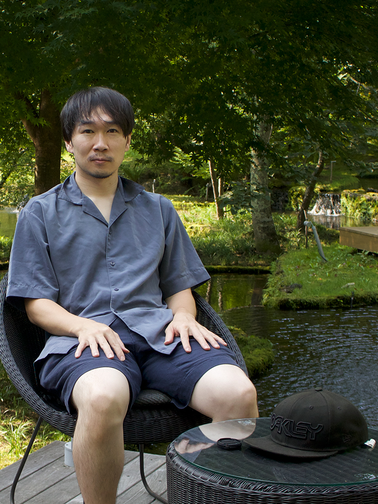 A man sits outdoors on a wicker chair by a pond, with a cap and small object on a glass table beside him. The setting is lush and green, suggesting a peaceful natural environment.