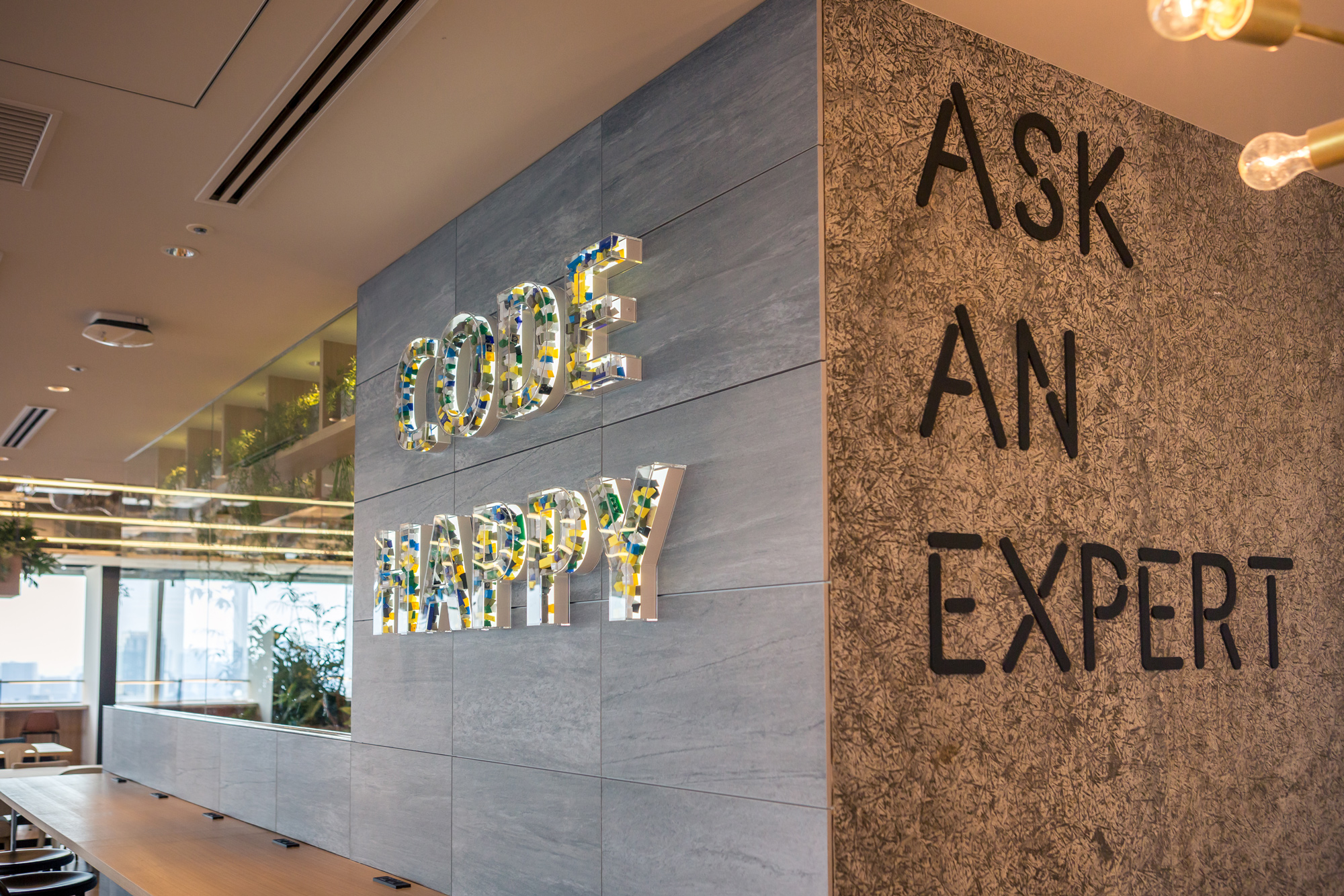 A modern office workspace featuring a wall with the phrases 'CODE HAPPY' in colorful, three-dimensional letters and 'ASK AN EXPERT' in bold black letters. The area includes seating and natural light, conveying a collaborative and inviting environment.