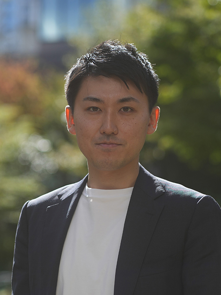 A portrait of a man standing outdoors wearing a dark blazer and white shirt, with greenery and buildings blurred in the background.