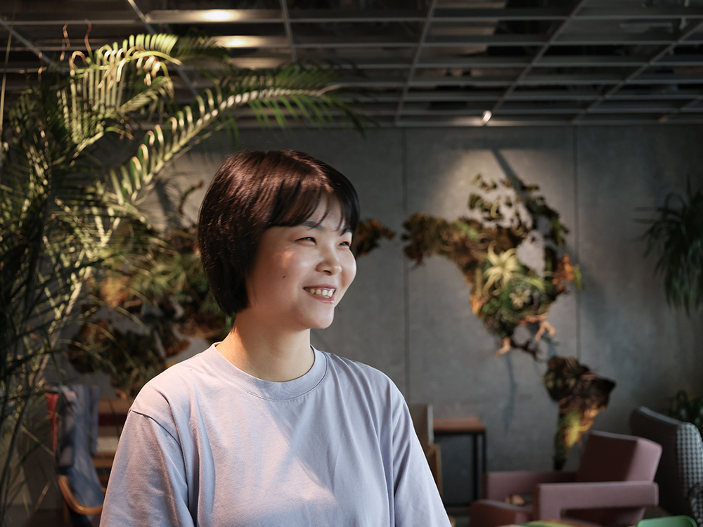 A person smiling indoors with a green wall and plants in the background, featuring a modern and natural office environment.
