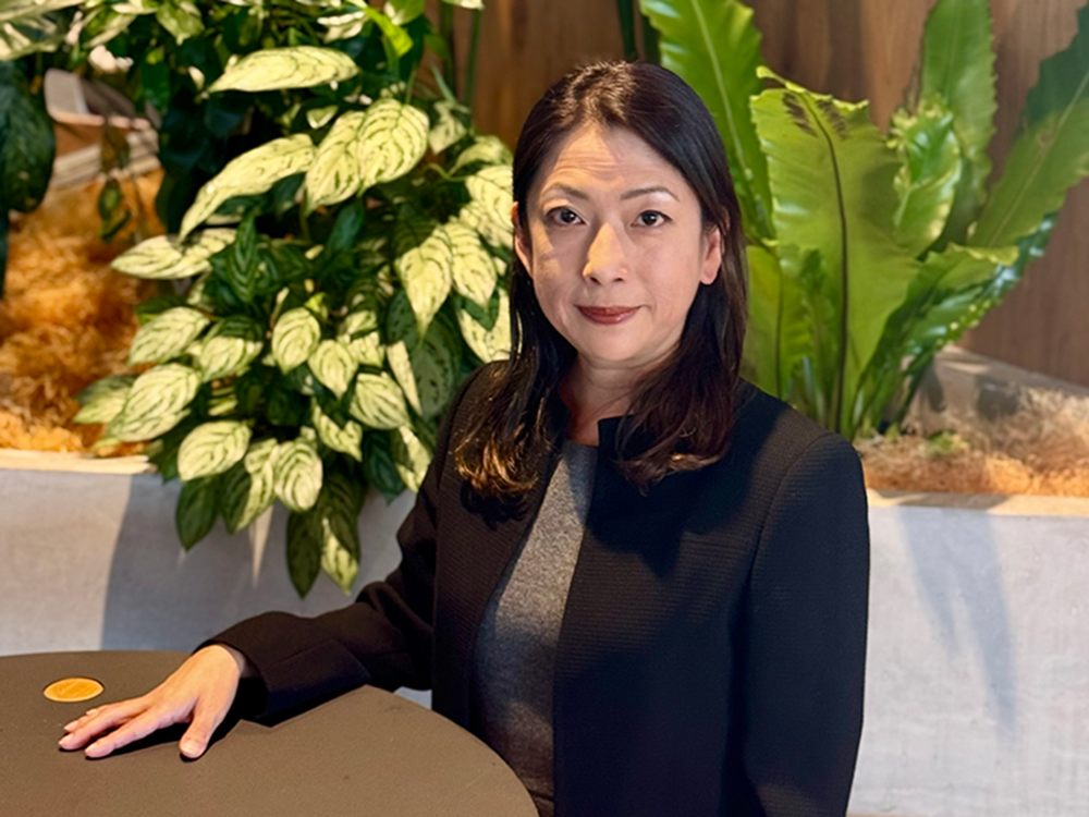 A woman in professional attire sitting at a table with a backdrop of lush indoor plants.