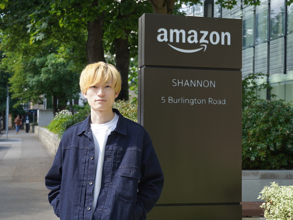 A person stands in front of the Amazon Shannon office sign at 5 Burlington Road, with trees and modern buildings in the background.