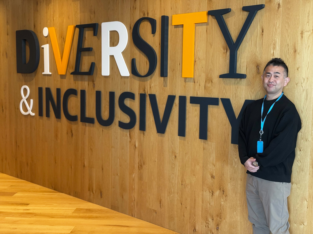 A person stands next to a large wall sign that reads 'Diversity & Inclusivity' in an office setting with wood panel walls and flooring.
