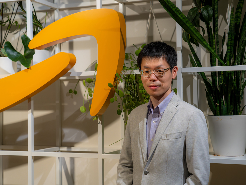 A man wearing a light-colored suit stands in front of a large yellow Amazon logo and a display of indoor plants.