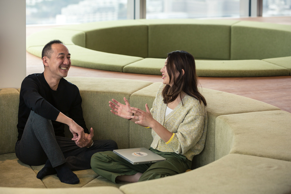 Two people sitting on a circular sofa in a modern office lounge, smiling and having a friendly conversation. One person holds a laptop with an AWS logo.