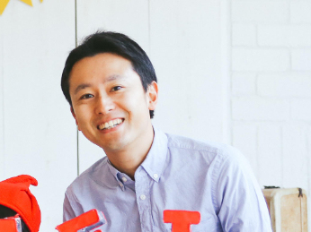A smiling man wearing a light blue shirt, indoors, with a white brick wall in the background.