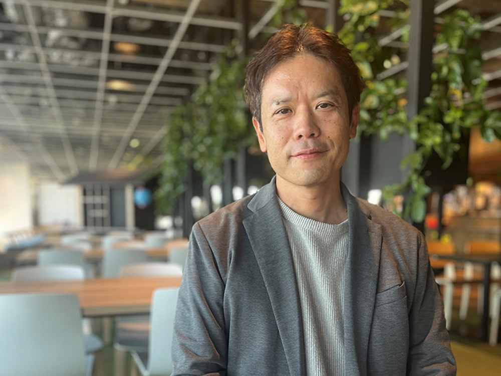 A man wearing a casual blazer is standing in a modern office space with a background of tables, chairs, and green plants.