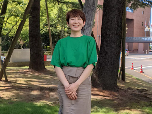 A woman wearing a green shirt and beige skirt is standing outdoors on grass, smiling, with large trees and a road in the background.