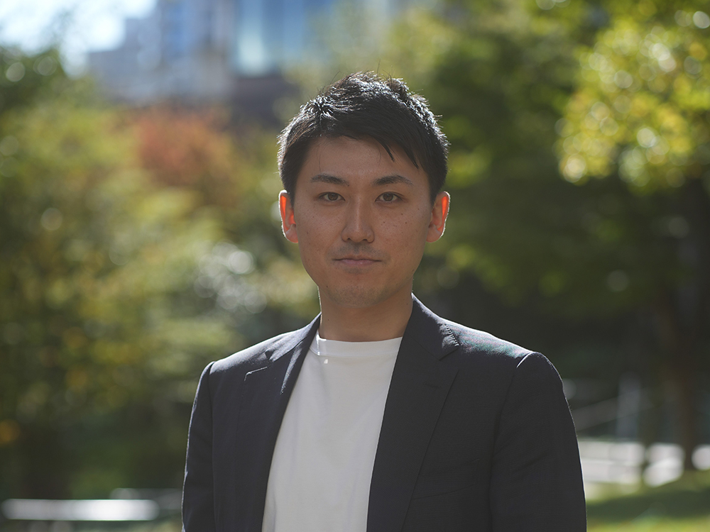 A man wearing a suit jacket stands outdoors in natural light, with greenery and blurred buildings in the background.
