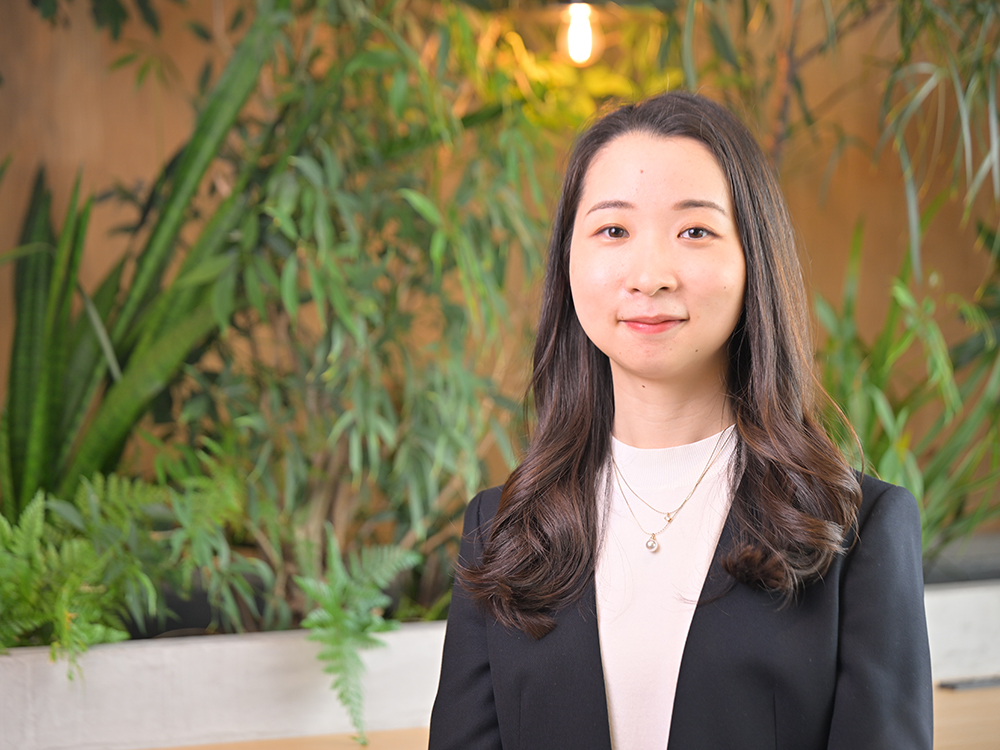 A professional portrait of a woman standing in front of a background with various indoor plants, creating a fresh and natural office atmosphere.