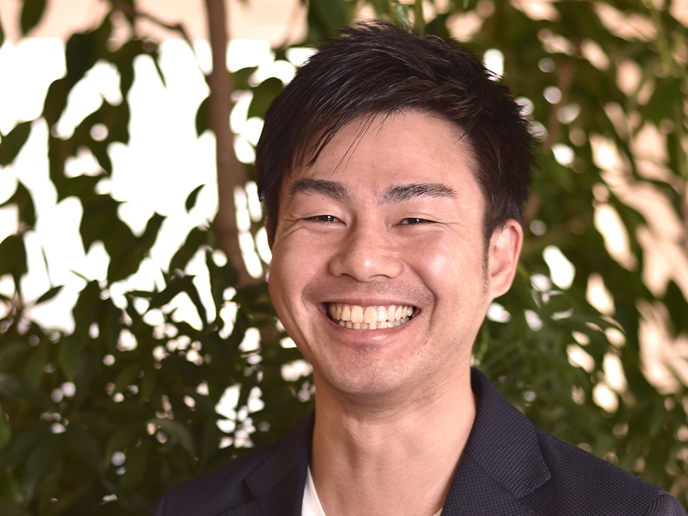 A portrait of a smiling man standing in front of indoor plants, with a warm and inviting expression.