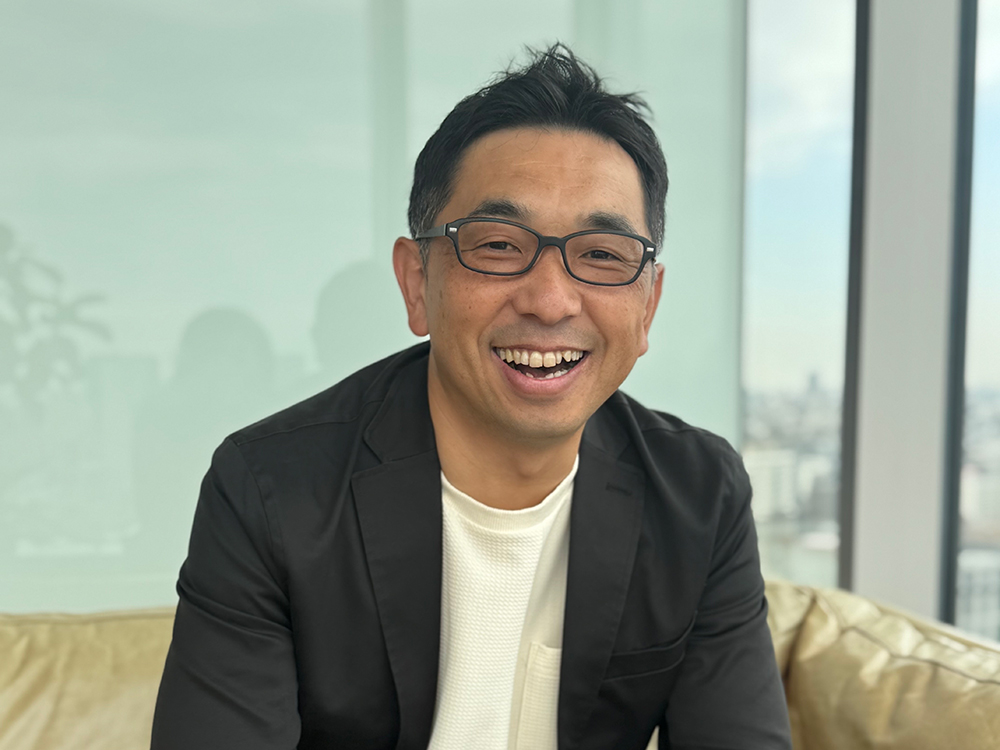 A smiling man wearing glasses and a black blazer sitting indoors with a city view in the background.