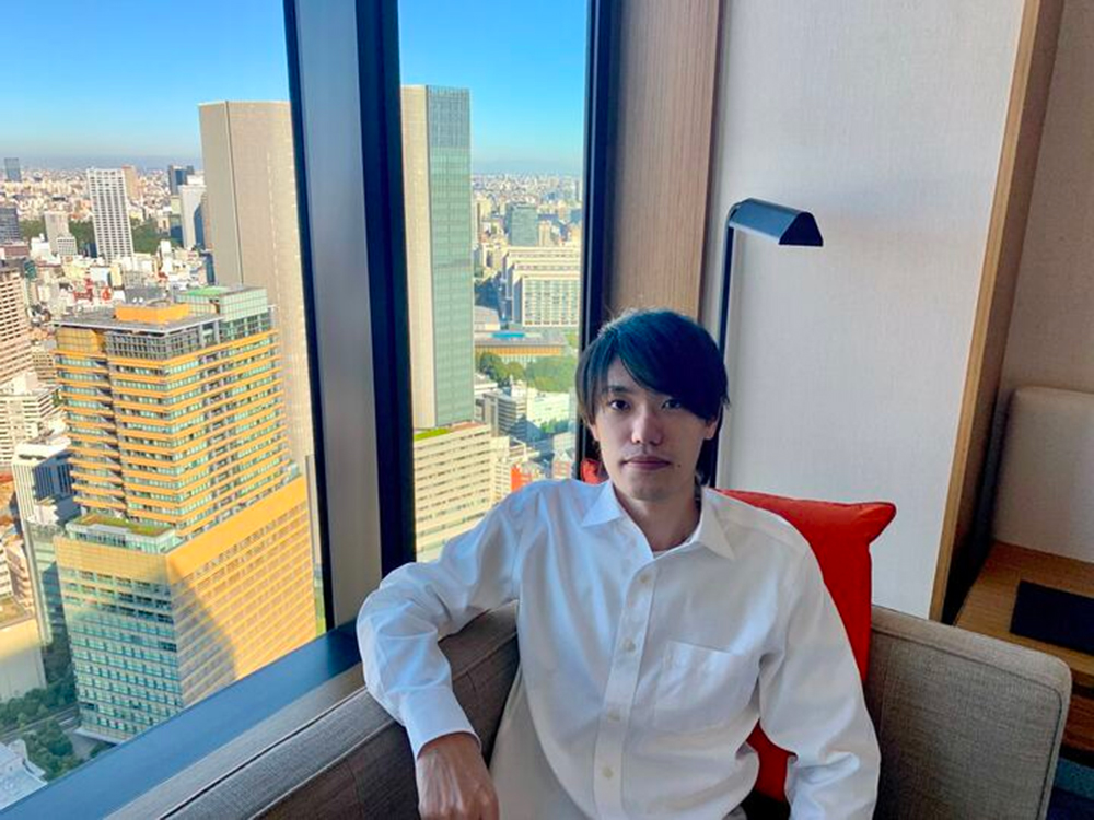 A man in a white shirt sits by a window in a modern room, with a cityscape featuring tall buildings visible outside during the daytime.