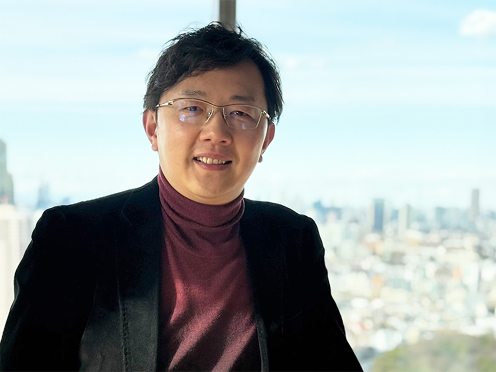 A man wearing glasses and a dark blazer stands in front of a large window, with a bright cityscape blurred in the background.